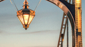 Kaiser-Wilhelm-Brücke bei Sonnenuntergang, beleuchtet, mit Laterne in der Mitte, umgeben von blauem Himmel.