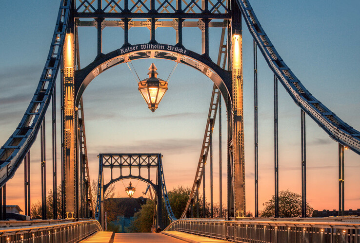 Kaiser-Wilhelm-Brücke bei Sonnenuntergang, beleuchtet, mit Laterne in der Mitte, umgeben von blauem Himmel.