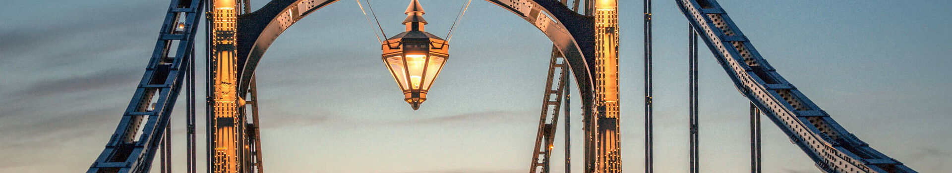 Kaiser-Wilhelm-Brücke bei Sonnenuntergang, beleuchtet, mit Laterne in der Mitte, umgeben von blauem Himmel.