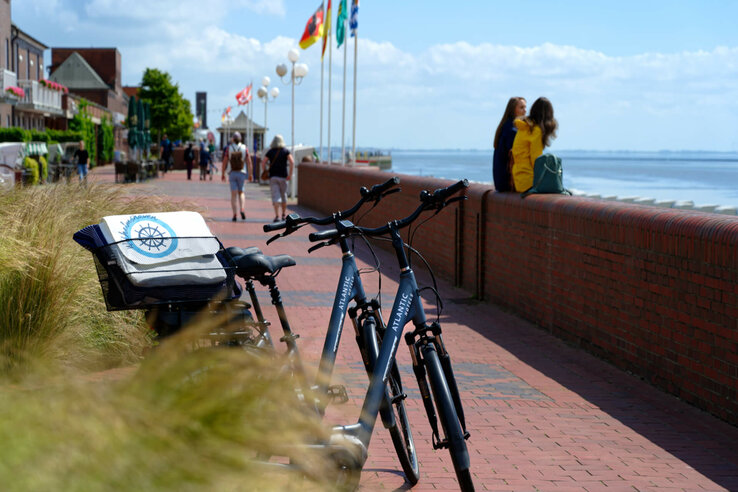Fahrräder an einer Uferpromenade mit Blick aufs Meer, Menschen spazieren, Flaggen wehen im Wind.