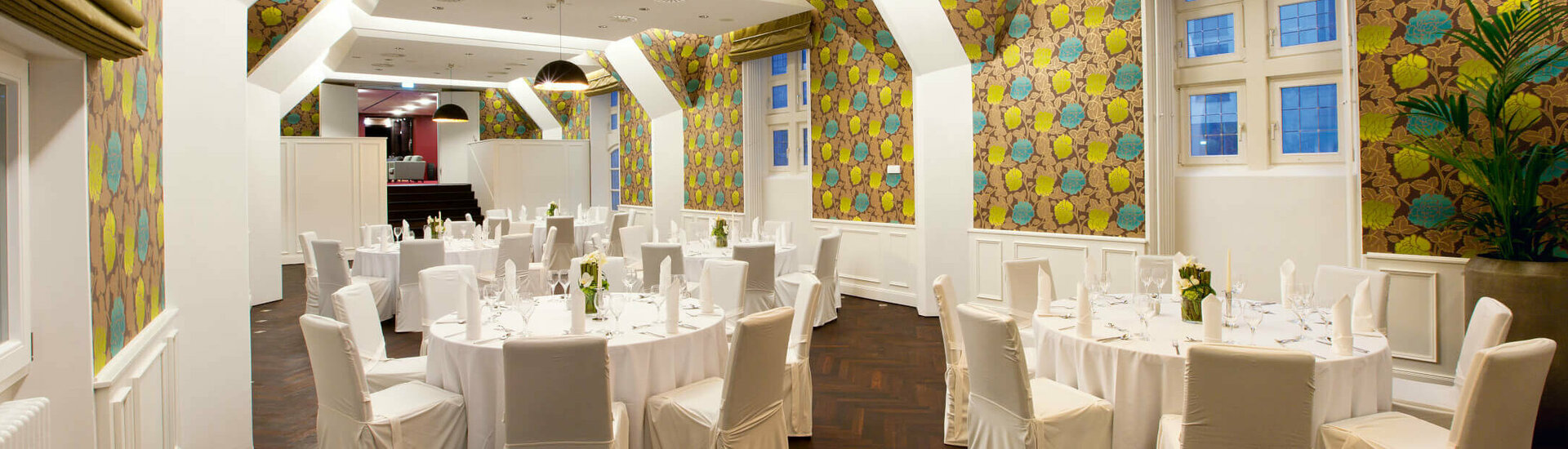 Elegant banquet hall in the ATLANTIC Grand Hotel Bremen with round, white-clothed tables and colorful wallpaper.