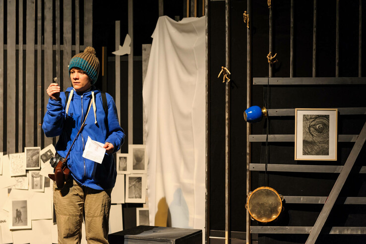 Person in blue jacket and cap holding paper, surrounded by works of art and props in a room. Schnürschuh Theater Bremen