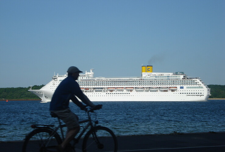 A cyclist rides along the sea while a large cruise ship passes by in the background.