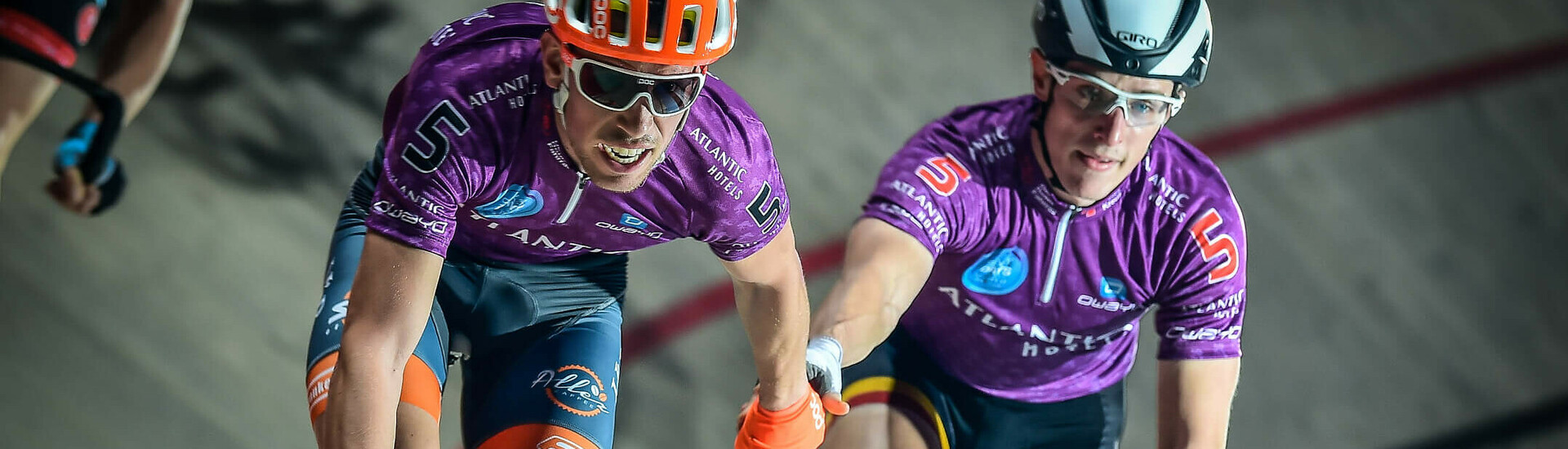 Zwei Radfahrer in lila Trikots fahren Seite an Seite auf einer Bahn und reichen sich die Hand.