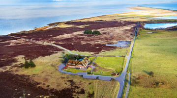 Luftaufnahme eines Hotels an der Küste, umgeben von grünen Feldern und Heidekraut, mit Blick auf das Meer im Hintergrund.