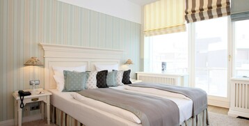 Bright hotel room with double bed, striped wallpaper, several pillows, bedside lamps and large windows with blinds.