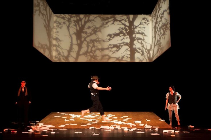Three people on a stage with paper leaves, tree projection in the background. © bremer shakespeare company