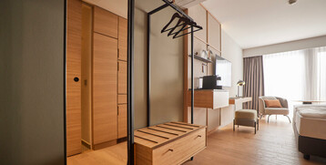 Modernes Hotelzimmer mit Holzfußboden, Doppelbett, Schreibtisch, Sessel und großem Fenster mit Vorhängen.