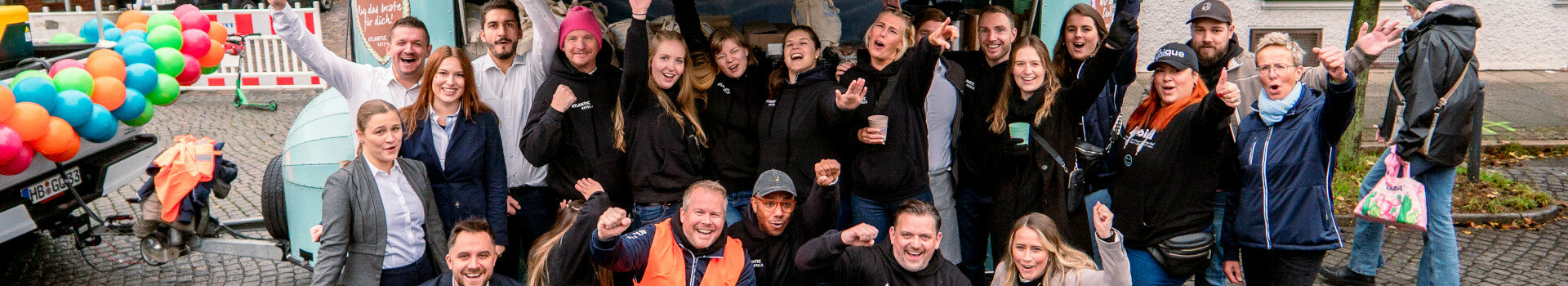 Gruppe fröhlicher Menschen in Freizeitkleidung posiert draußen vor einem bunt geschmückten Anhänger.