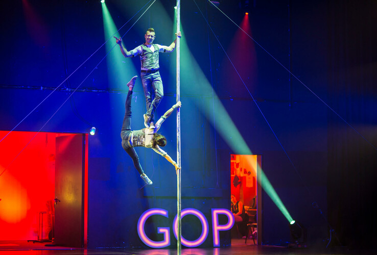 Zwei Akrobaten führen eine Show an einer vertikalen Stange auf, beleuchtet von bunten Scheinwerfern.