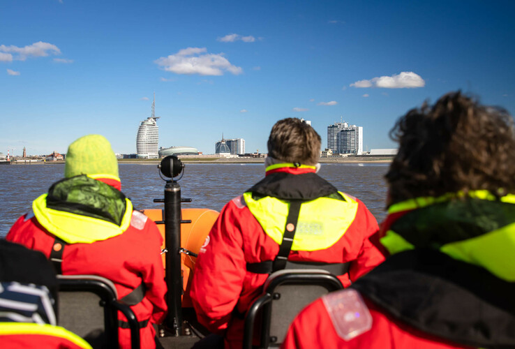 Personen in roten Jacken auf einem Boot, blicken auf das ATLANTIC Hotel Sail City in Bremerhaven bei sonnigem Wetter.