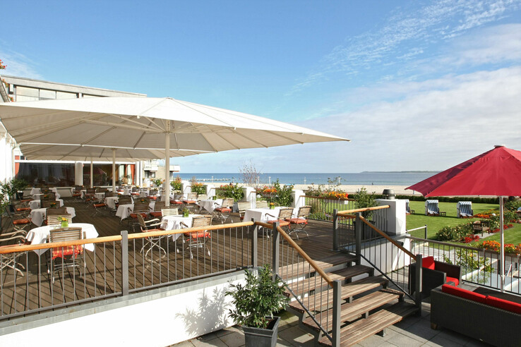 Hotelterrasse mit Meerblick, weißen Tischen, Holzstühlen und großen Sonnenschirmen, umgeben von Blumen und grüner Wiese.