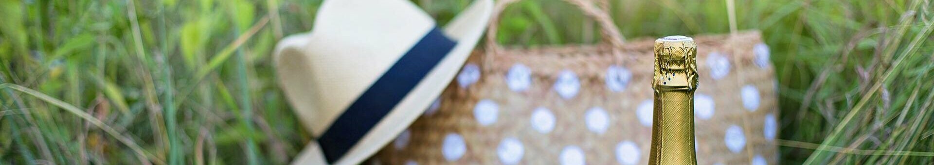 Picnic in the countryside with a bottle of champagne, two glasses, straw hat and polka dot bag in the grass.