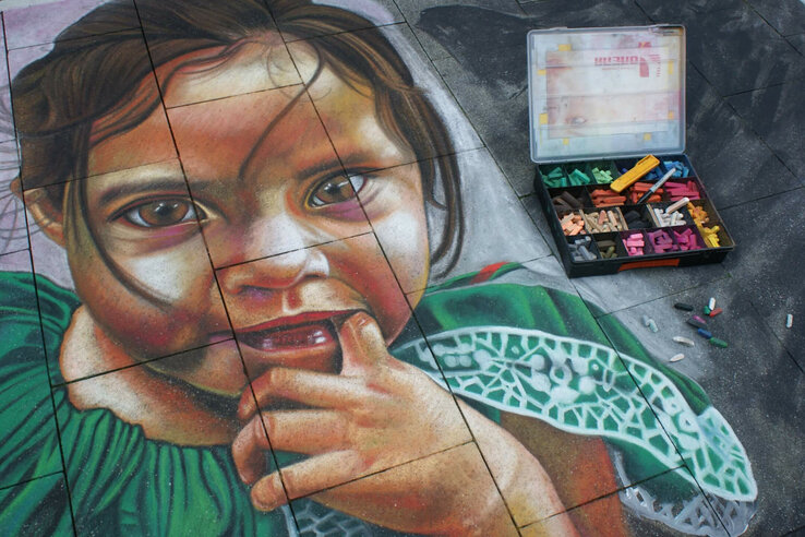 Street art: children's faces in chalk on paving stones, next to colorful pieces of chalk in an open box.