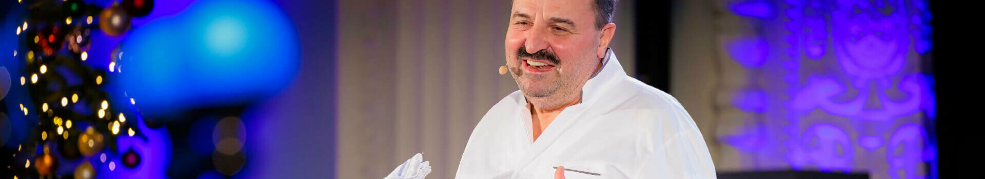 Ein Koch im weißen Outfit präsentiert auf einer Bühne, im Hintergrund ein dekorierter Weihnachtsbaum.