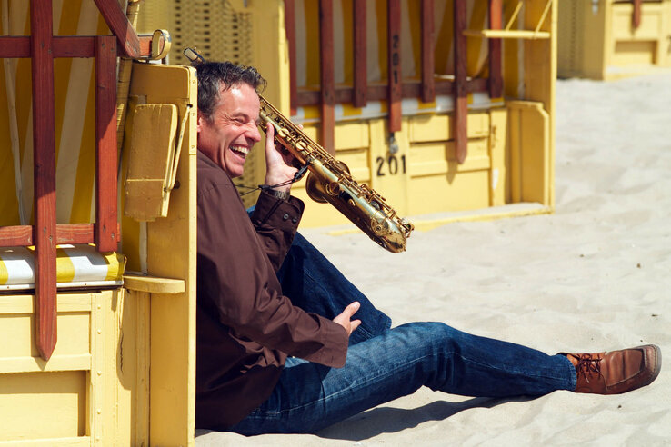 Mann mit Saxophon sitzt lachend am Strand vor gelbem Strandkorb.