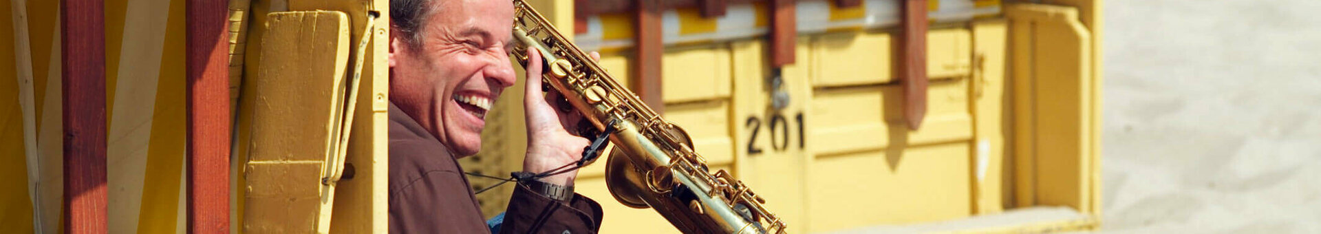 Mann mit Saxophon sitzt lachend am Strand vor gelbem Strandkorb.