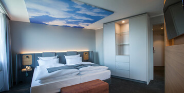Modernes Hotelzimmer mit Doppelbett, Himmelmotiv an der Decke, Schrank, Flachbildfernseher und Sitzbank im ATLANTIC Hotel.