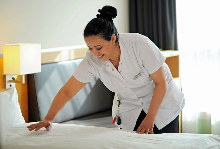 Hotelmitarbeiterin richtet lächelnd ein Bett her, helle und freundliche Atmosphäre.