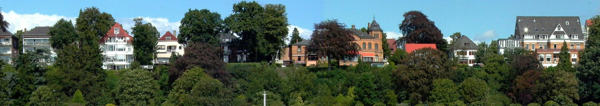 Blick auf eine grüne Flussuferpromenade mit historischen Gebäuden im Hintergrund, unter blauem Himmel.