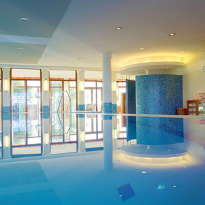 Pool im ATLANTIC Hotel Wilhelmshaven Helles Hallenbad mit großen Fenstern, blau gefliesten Säulen und Liegestühlen im Hintergrund.