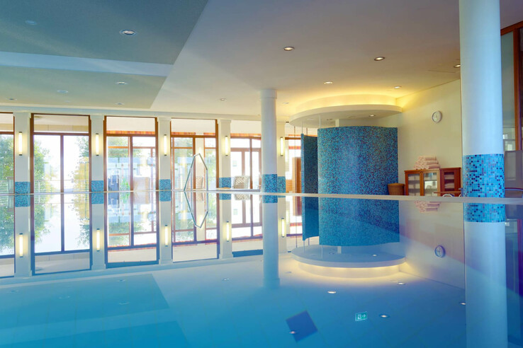 Helles Hallenbad mit großen Fenstern, blau gefliesten Säulen und Liegestühlen im Hintergrund.
