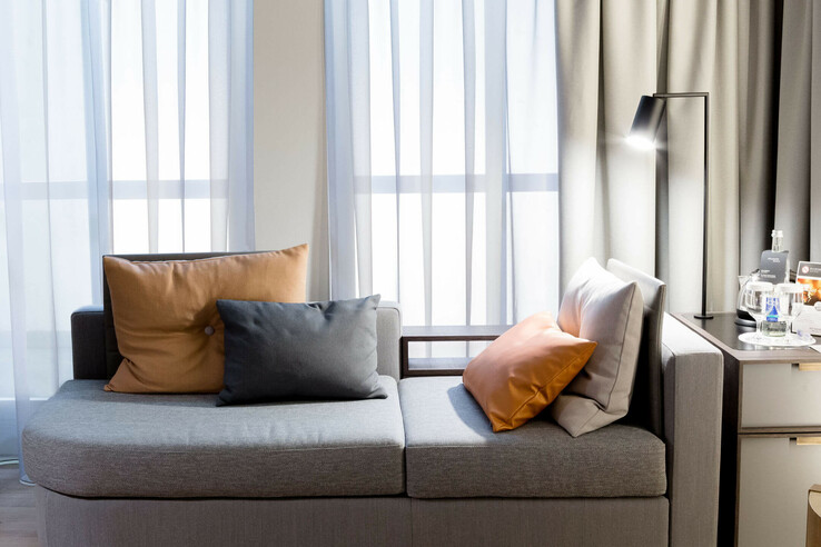 Gray sofa with orange cushions, floor lamp and side table with drinks in front of light-colored curtains in the ATLANTIC Grand Hotel Bremen.