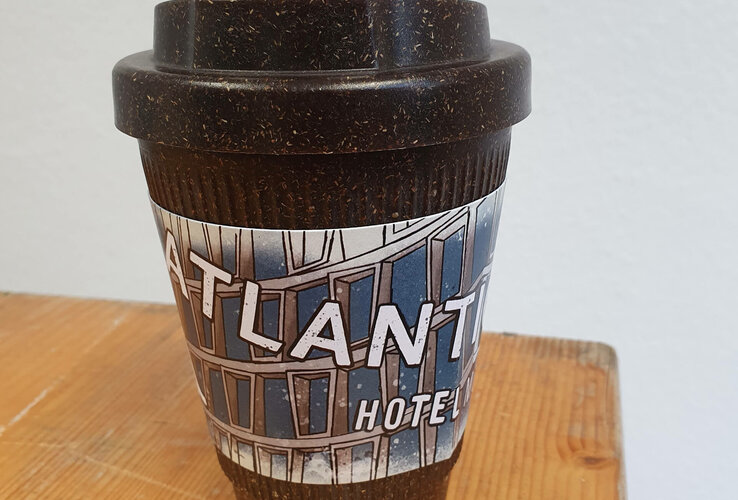Reusable coffee mug with ATLANTIC Hotel Münster logo on wooden table.