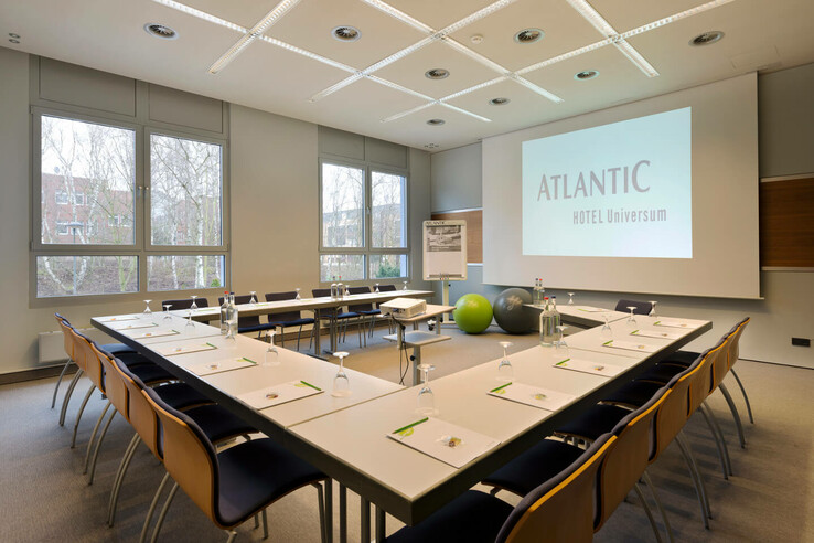 Heller Konferenzraum im ATLANTIC Hotel Universum mit U-förmiger Tischanordnung, Leinwand und Präsentationsmaterial.