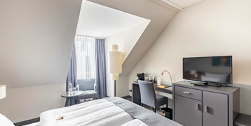Helles Hotelzimmer mit Doppelbett, Schreibtisch, Fernseher, Sessel und großem Fenster im ATLANTIC Hotel Vegesack.
