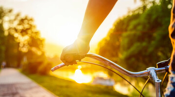 Person hält Fahrradlenker bei Sonnenuntergang, entlang eines Flusses mit grünen Bäumen im Hintergrund.