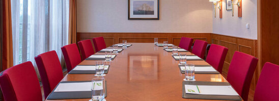 Konferenzraum mit langem Holztisch, roten Stühlen, Notizblöcken und Wasser, beleuchtet durch elegante Deckenlampen.