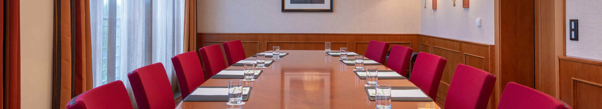 Konferenzraum mit langem Holztisch, roten Stühlen, Notizblöcken und Wasser, beleuchtet durch elegante Deckenlampen.