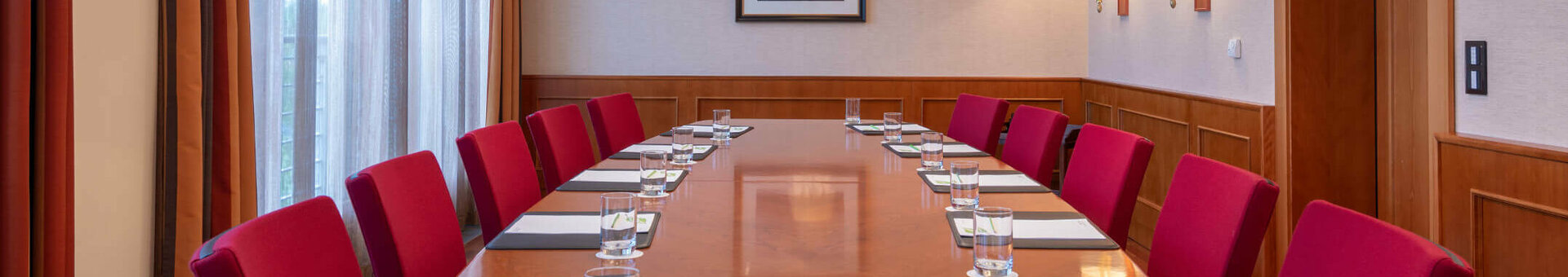 Conference room with a long wooden table, red chairs, notepads and water, illuminated by elegant ceiling lamps.