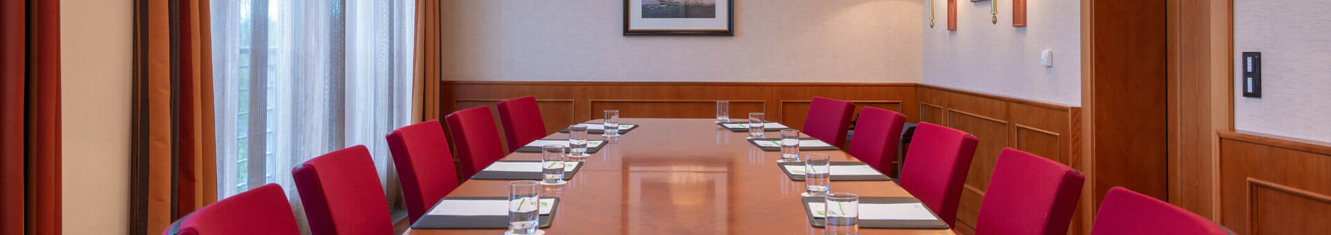 Konferenzraum mit langem Holztisch, roten Stühlen, Notizblöcken und Wasser, beleuchtet durch elegante Deckenlampen.