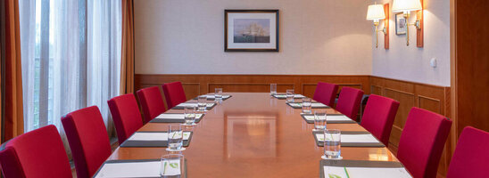 Conference room with a long wooden table, red chairs, notepads and water, illuminated by elegant ceiling lamps.