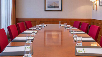 Konferenzraum mit langem Holztisch, roten Stühlen, Notizblöcken und Wasser, beleuchtet durch elegante Deckenlampen.