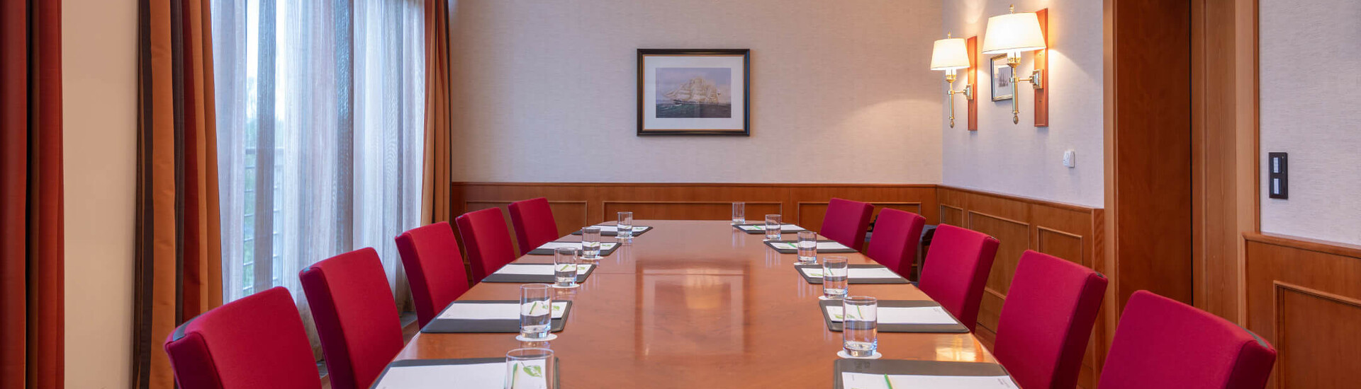 Tagungsraum Münster | ATLANTIC Hotel Wilhelmshaven Konferenzraum mit langem Holztisch, roten Stühlen, Notizblöcken und Wasser, beleuchtet durch elegante Deckenlampen.