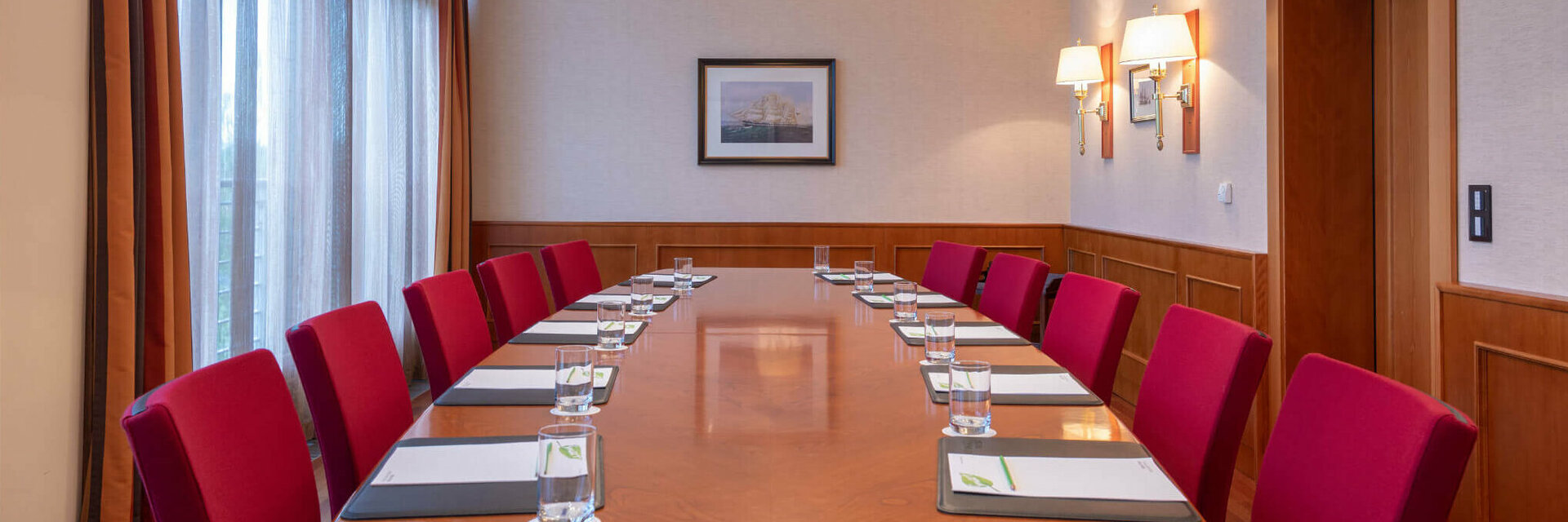 Konferenzraum mit langem Holztisch, roten Stühlen, Notizblöcken und Wasser, beleuchtet durch elegante Deckenlampen.