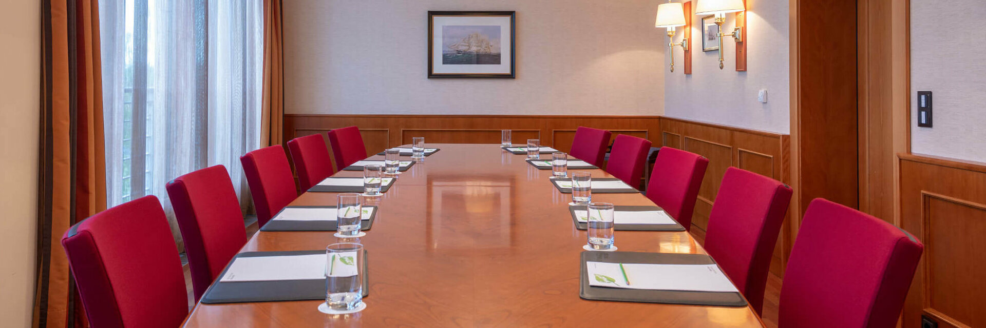 Conference room with a long wooden table, red chairs, notepads and water, illuminated by elegant ceiling lamps.