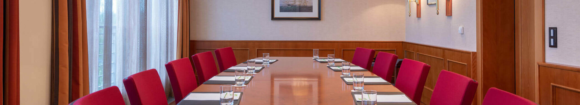 Conference room with a long wooden table, red chairs, notepads and water, illuminated by elegant ceiling lamps.