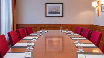 Conference room with a long wooden table, red chairs, notepads and water, illuminated by elegant ceiling lamps.