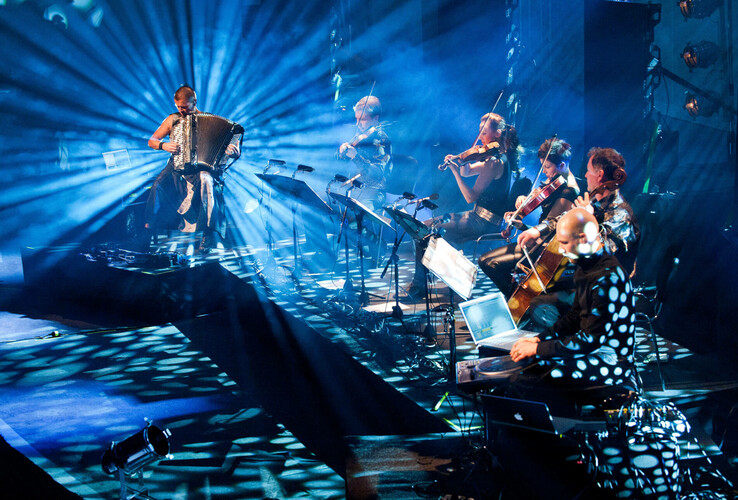 Musiker auf Bühne mit Akkordeon und Streichern, umgeben von blauem Licht und Strahlen.