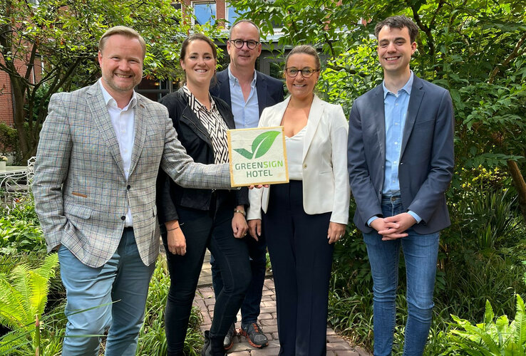 Pressemitteilung | ATLANTIC Hotels Gruppe GreenSign zertifiziert Eine Gruppe von fünf Personen steht lächelnd im Garten eines Hotels und hält ein "Greensign Hotel"-Schild.