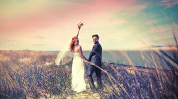 Brautpaar am Strand mit Blick aufs Meer, Frau hebt Blumenstrauß, romantische Atmosphäre.