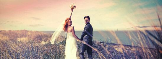 Brautpaar am Strand mit Blick aufs Meer, Frau hebt Blumenstrauß, romantische Atmosphäre.