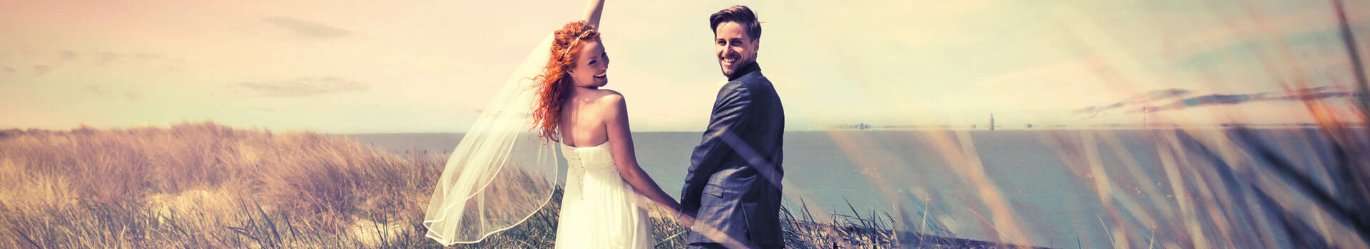 Brautpaar am Strand mit Blick aufs Meer, Frau hebt Blumenstrauß, romantische Atmosphäre.