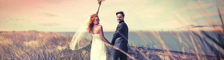 Brautpaar am Strand mit Blick aufs Meer, Frau hebt Blumenstrauß, romantische Atmosphäre.