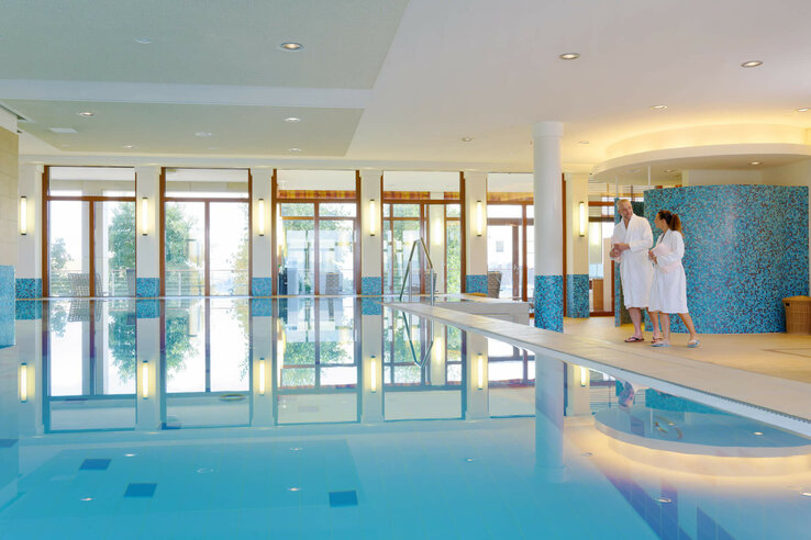 Bright indoor pool with blue mosaic, large windows and two people in bathrobes at the edge of the pool.