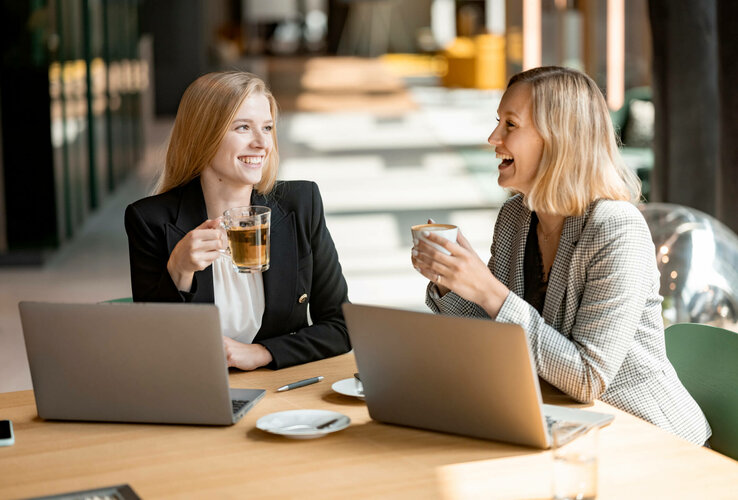 Two women smile and drink coffee in a modern hotel lounge area with laptops on the table.